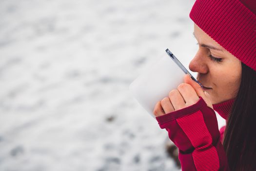 Beautiful young caucasian woman with brown hair, wearing a red jacket and a hat outside in the forest when it's snowing. Woman walking around a snowy forest. Woman holding a to go cup with tea. 