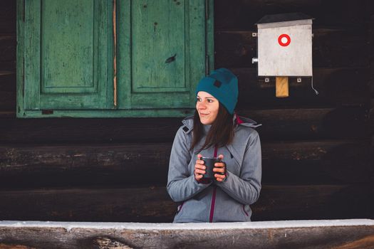 Beautiful young caucasian woman with brown hair, wearing a red jacket and a hat outside in the forest when it's snowing. Woman walking around a snowy forest. Woman holding a to go cup with tea. 