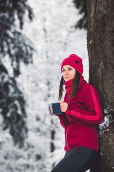 Beautiful young caucasian woman with brown hair, wearing a red jacket and a hat outside in the forest when it's snowing. Woman walking around a snowy forest. Woman holding a to go cup with tea. 