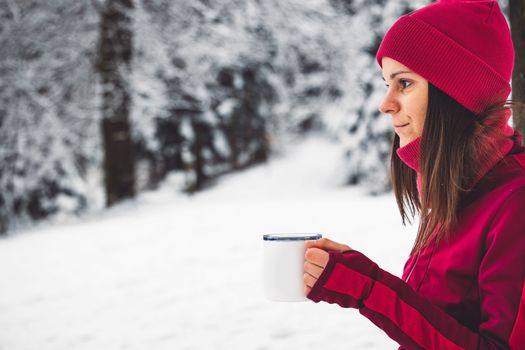 Beautiful young caucasian woman with brown hair, wearing a red jacket and a hat outside in the forest when it's snowing. Woman walking around a snowy forest. Woman holding a to go cup with tea. 