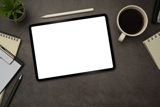 Flat lay digital tablet with white empty display, stylus pen, coffee cup and notepad on black slate texture background.