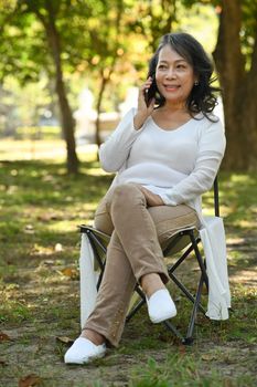 Full length of middle age woman in casual clothes talking on mobile phone while sitting in the park surrounded nature view.