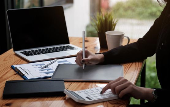 Business woman or accountant hand using calculator, working on taplet and laptop computer with documentary data graph paper on the table at modern home office. Online marketing concept..