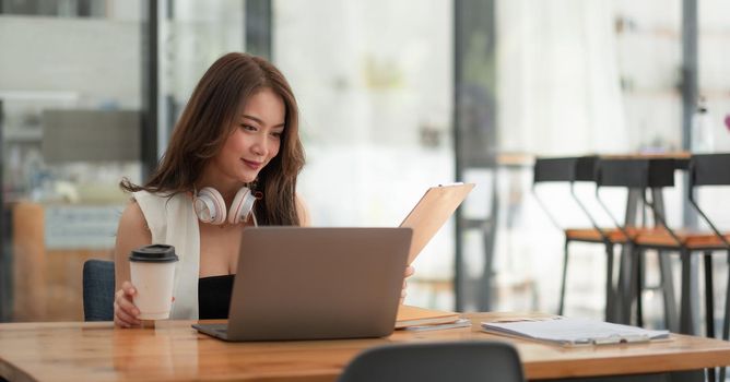 Smiling girl student wear wireless headphone study online with teacher, happy young asian woman learn language listen lecture watch webinar write notes look at laptop sit in cafe, distant education