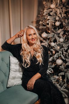 A girl in an evening dress is sitting on the sofa near the Christmas tree.