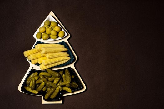 Dish in the form of a Christmas tree with pickled vegetables on a brown background.A plate of cucumbers, corn and olives at Christmas