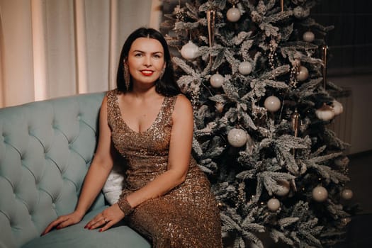 A girl in an evening dress is sitting on the sofa near the Christmas tree.