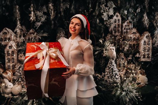 a girl in a Santa hat with a big Christmas gift in her hands on a fairy-tale island.Smiling woman in white clothes on the background of Christmas trees and small houses.