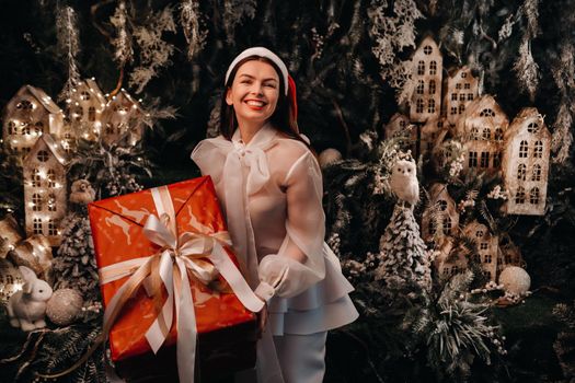 a girl in a Santa hat with a big Christmas gift in her hands on a fairy-tale island.Smiling woman in white clothes on the background of Christmas trees and small houses.