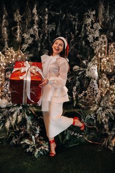 a girl in a Santa hat with a big Christmas gift in her hands on a fairy-tale island.Smiling woman in white clothes on the background of Christmas trees and small houses.