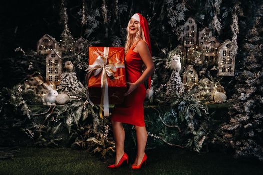 a girl in a Santa hat with a big Christmas gift in her hands on a fabulous Christmas background.A smiling woman in a red dress on the background of Christmas trees and small houses.