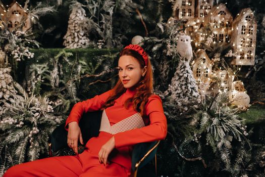 a girl in a red suit is sitting on a chair near the Christmas background. a woman in red on the background of Christmas trees and small Christmas houses.