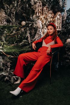 a girl in a red suit is sitting on a chair near the Christmas background. a woman in red on the background of Christmas trees and small Christmas houses.