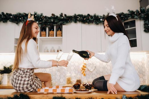 two girls in a cozy home environment in the kitchen pour champagne for Christmas. Smiling girls drink champagne on a festive evening.