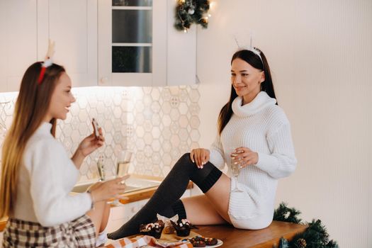 two girls in a cozy home environment in the kitchen with champagne in their hands for Christmas. Smiling girls drink champagne on a festive evening.