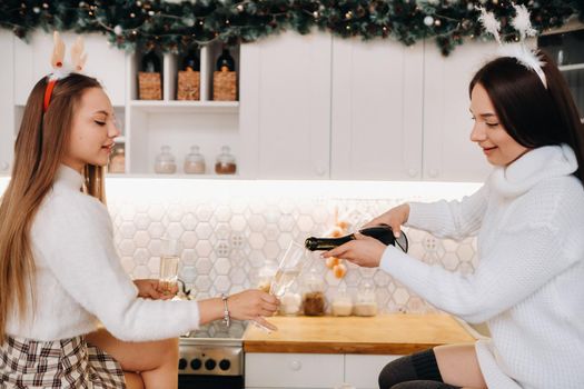 two girls in a cozy home environment in the kitchen pour champagne for Christmas. Smiling girls drink champagne on a festive evening.