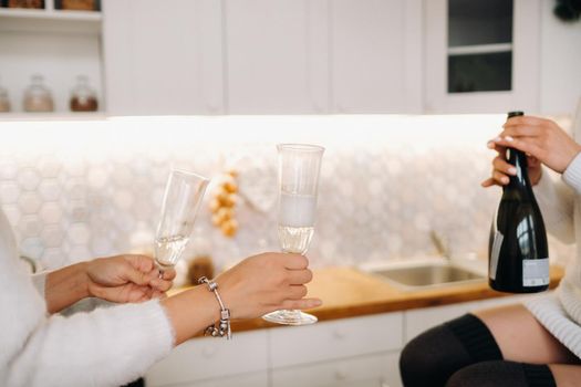 two girls in a cozy home environment in the kitchen pour champagne for Christmas. Smiling girls drink champagne on a festive evening.