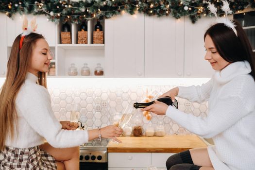 two girls in a cozy home environment in the kitchen pour champagne for Christmas. Smiling girls drink champagne on a festive evening.
