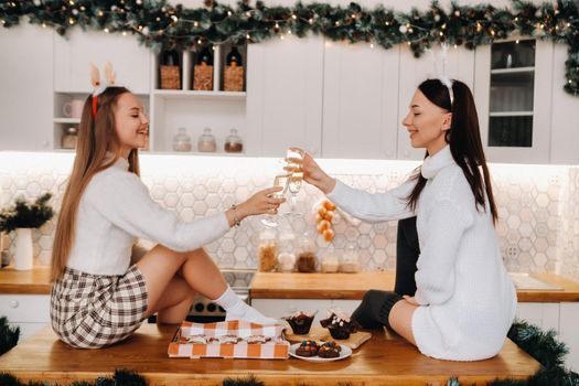 two girls in a cozy home environment in the kitchen with champagne in their hands for Christmas. Smiling girls drink champagne on a festive evening.