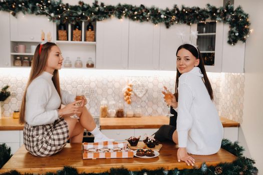 two girls in a cozy home environment in the kitchen with champagne in their hands for Christmas. Smiling girls drink champagne on a festive evening.