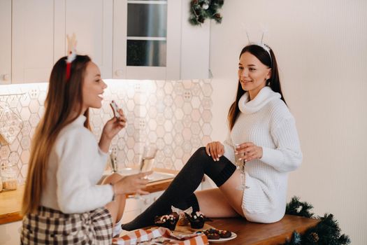 two girls in a cozy home environment in the kitchen with champagne in their hands for Christmas. Smiling girls drink champagne on a festive evening.