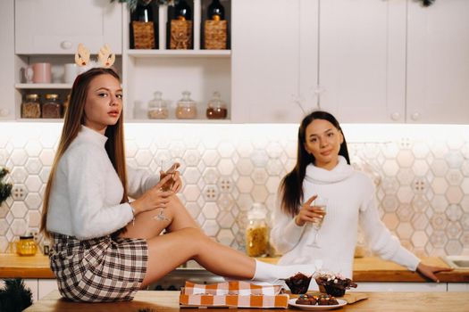 two girls in a cozy home environment in the kitchen with champagne in their hands for Christmas. Smiling girls drink champagne on a festive evening.