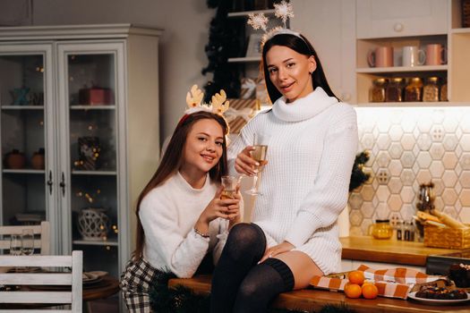 two girls in a cozy home environment in the kitchen with champagne in their hands for Christmas. Smiling girls drink champagne on a festive evening.