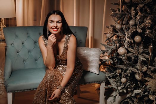 A girl in an evening dress is sitting on the sofa near the Christmas tree.