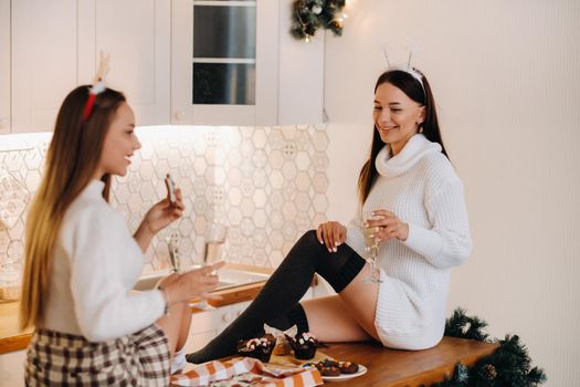 two girls in a cozy home environment in the kitchen with champagne in their hands for Christmas. Smiling girls drink champagne on a festive evening.