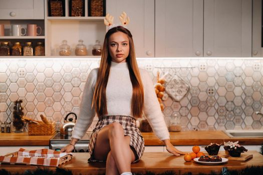 A girl with reindeer horns at Christmas sits on the kitchen table and smiles.Woman in new year's in the kitchen.