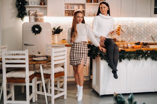 two girls in a cozy home environment in the kitchen with champagne in their hands for Christmas. Smiling girls drink champagne on a festive evening.