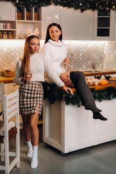 two girls in a cozy home environment in the kitchen with champagne in their hands for Christmas. Smiling girls drink champagne on a festive evening.