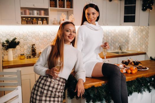 two girls in a cozy home environment in the kitchen with champagne in their hands for Christmas. Smiling girls drink champagne on a festive evening.