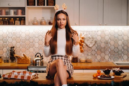 A girl with reindeer horns at Christmas sits on the kitchen table and smiles.Woman in new year's in the kitchen.