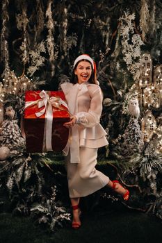 a girl in a Santa hat with a big Christmas gift in her hands on a fairy-tale island.Smiling woman in white clothes on the background of Christmas trees and small houses.