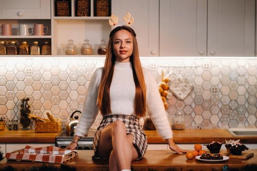 A girl with reindeer horns at Christmas sits on the kitchen table and smiles.Woman in new year's in the kitchen.