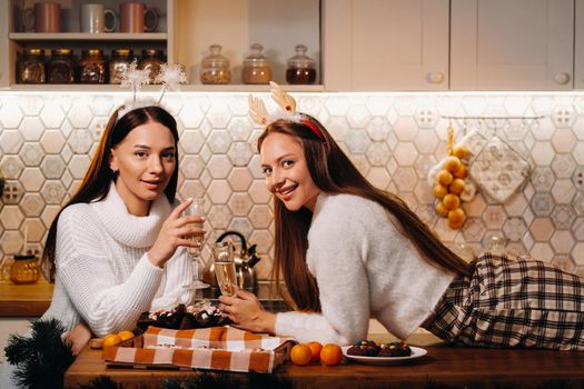 two girls in a cozy home environment with champagne in their hands at Christmas. Smiling girls drink champagne on a festive evening.