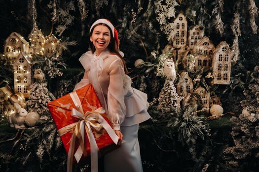 a girl in a Santa hat with a big Christmas gift in her hands on a fairy-tale island.Smiling woman in white clothes on the background of Christmas trees and small houses.