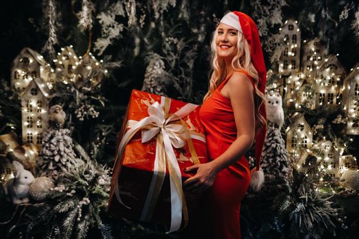 a girl in a Santa hat with a big Christmas gift in her hands on a fabulous Christmas background.A smiling woman in a red dress on the background of Christmas trees and small houses.