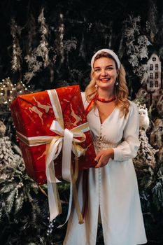 a girl in a white dress with a large Christmas gift in her hands on a fairy-tale background.Smiling woman in white clothes on the background of Christmas trees and small houses.