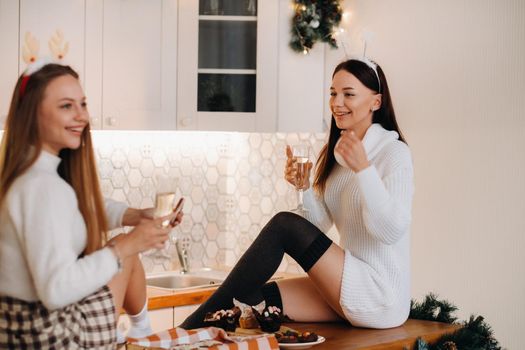 two girls in a cozy home environment in the kitchen with champagne in their hands for Christmas. Smiling girls drink champagne on a festive evening.