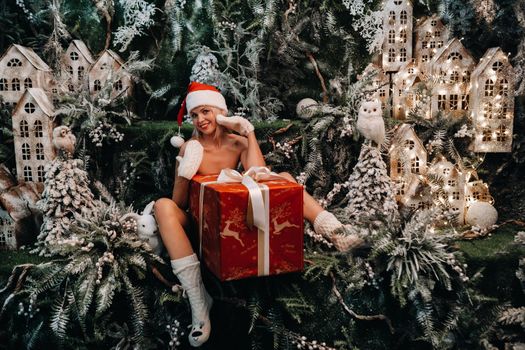 a girl in a Santa hat with a big Christmas gift in her hands on a fabulous Christmas background.Smiling woman in a bathing suit on the background of Christmas trees and small houses.