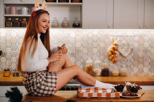 A girl on Christmas day sits on the kitchen table and holds a glass of champagne in her hands.A woman on New year's eve in the kitchen with champagne.