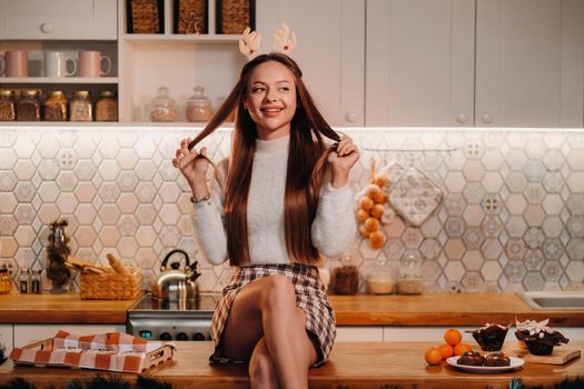 A girl with reindeer horns at Christmas sits on the kitchen table and smiles.Woman in new year's in the kitchen.