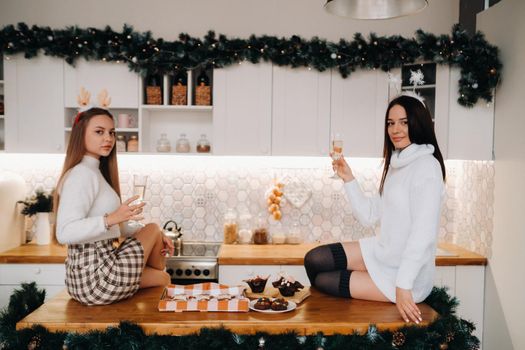 two girls in a cozy home environment in the kitchen with champagne in their hands for Christmas. Smiling girls drink champagne on a festive evening.