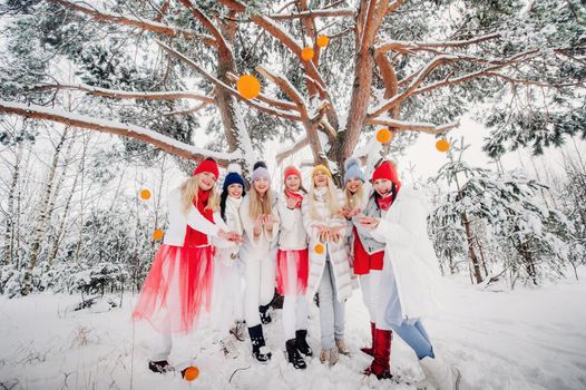 A large group of girls with tangerines are standing in the winter forest.Girls in red and white clothes with fruit in a snow-covered forest.