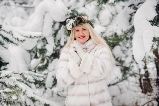 Portrait of a woman in a white fur coat in a cold winter forest. Girl with a wreath on her head in a snow-covered winter forest.