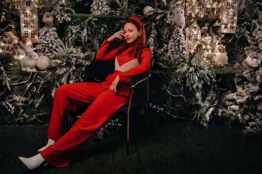 a girl in a red suit is sitting on a chair near the Christmas background. a woman in red on the background of Christmas trees and small Christmas houses.