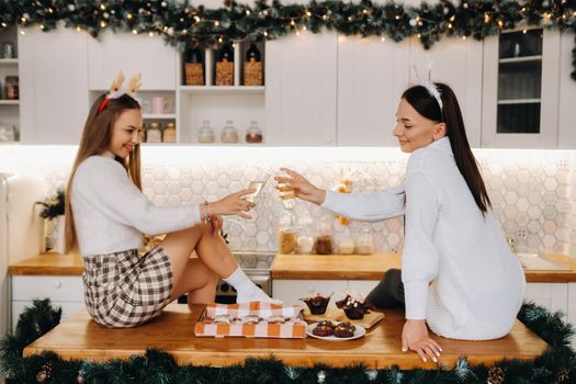 two girls in a cozy home environment in the kitchen with champagne in their hands for Christmas. Smiling girls drink champagne on a festive evening.