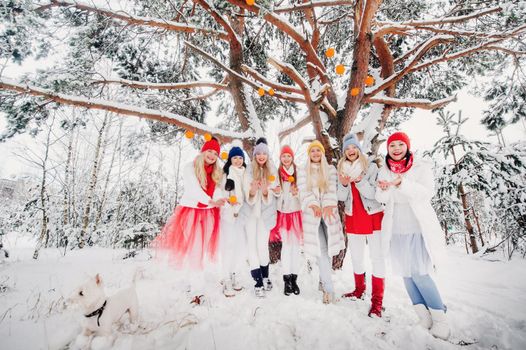 A large group of girls with tangerines are standing in the winter forest.Girls in red and white clothes with fruit in a snow-covered forest.